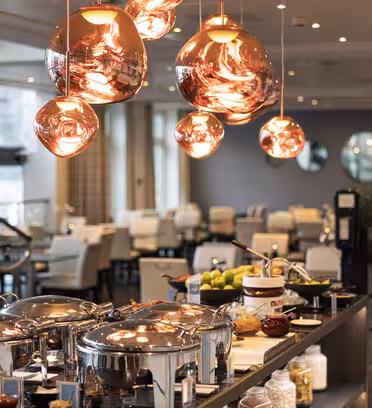 A modern restaurant buffet featuring copper pendant lights, an array of lidded serving dishes, fruits, and condiments on a long counter. In the background, white chairs and tables fill the dining area, creating a sleek and inviting atmosphere.