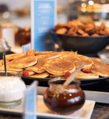 A plate of golden pancakes garnished with raspberries is displayed on a table. Next to it, there are small jars of honey and whipped cream. In the background, theres another bowl with assorted pastries slightly out of focus.
