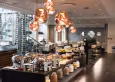 A modern restaurant interior with a buffet setup. Copper hanging lights illuminate chafing dishes, glass jars with grains, and bowls of fruit. In the background, tables and chairs are set up for dining, with large windows and a wine rack visible.