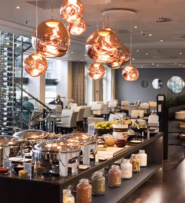 A modern restaurant interior with a buffet setup. Copper hanging lights illuminate chafing dishes, glass jars with grains, and bowls of fruit. In the background, tables and chairs are set up for dining, with large windows and a wine rack visible.
