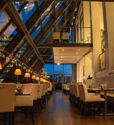 A modern restaurant interior with slanted glass walls offering a view of the city skyline at dusk. Tables are set with white chairs, and warm lamps create a cozy atmosphere. Holiday decorations hang along the wall.