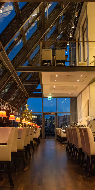 A modern restaurant interior with slanted glass walls offering a view of the city skyline at dusk. Tables are set with white chairs, and warm lamps create a cozy atmosphere. Holiday decorations hang along the wall.