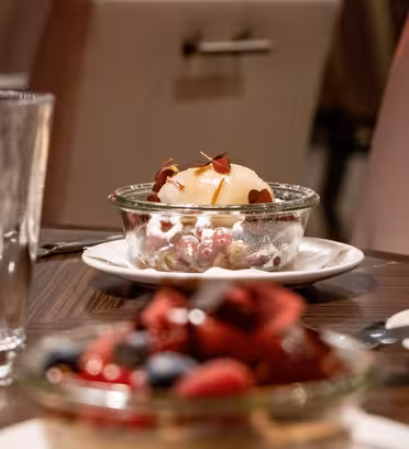 A dessert in a glass bowl sits on a table, topped with a scoop of vanilla ice cream and garnished with red microgreens. Blurred in the foreground is another dessert with mixed berries. A glass of water and cutlery are also visible.
