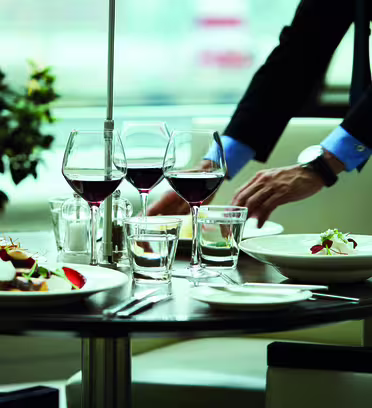 A table set in a restaurant with elegant dishes, including plates of salad and wine glasses. A person in formal attire adjusts the table settings. Background features a plant and soft lighting, creating a sophisticated ambiance.