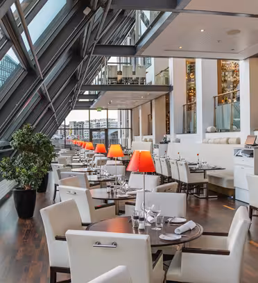 Modern restaurant interior with floor-to-ceiling windows and angled glass walls. White upholstered chairs surround dark wooden tables set with napkins and glassware. Warm-toned lamps and a large potted plant add a cozy ambiance.