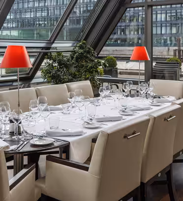 Elegant dining area with a large table set for ten, featuring white tablecloth, neatly arranged glassware, and silverware. Red lamps and green plants accent the space, with large glass windows offering a view of an urban exterior.