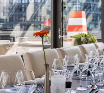 A restaurant table is set with empty wine glasses, water glasses, and white napkins. A single orange flower in a vase decorates the center, with large windows and a blurred cityscape in the background.