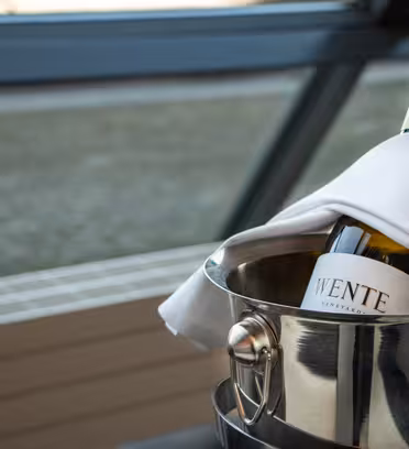 A bottle of white wine wrapped in a white napkin rests in a metal ice bucket. The wine label is visible. The background features blurred windows and a grid-patterned floor.