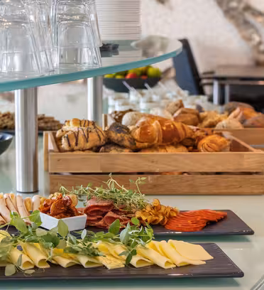 A buffet table with assorted cheeses, meats, fresh herbs, and a variety of breads in wooden crates. Stacked clear glasses are seen on a stand, and black chairs are visible in the background.