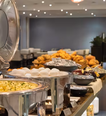 A buffet table is set with various dishes, including trays of scrambled eggs, boiled eggs, pastries, and baskets of bread. The table is elegantly arranged in a well-lit dining area with round mirrors reflecting the scene.