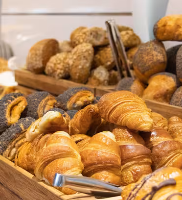 Assorted baked goods, including croissants, poppy seed rolls, and various breads, are displayed in wooden trays at a bakery or buffet setting. A pair of tongs is visible resting on the pastries.