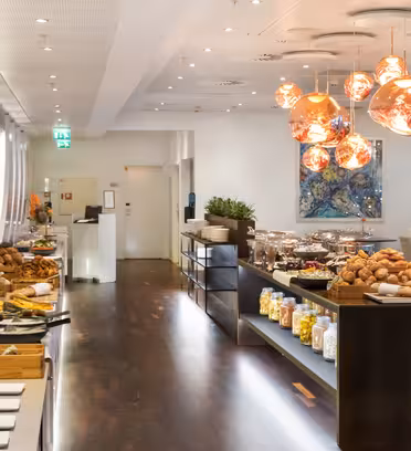 A spacious buffet area with a variety of breakfast items, including cereals, pastries, fruits, and condiments. Warm, decorative lighting hangs from the ceiling above the spread, giving a cozy ambiance to the modern, well-organized space.